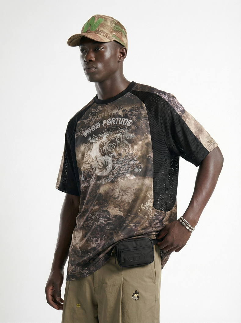 Model wearing a desert camo graphic mesh performance tee with black sleeves, khaki pants, and a camo cap