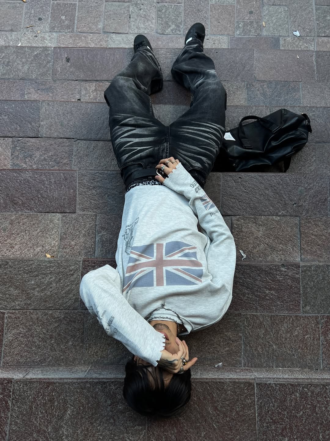 Person lying on stone pavement wearing a grey Union flag graphic long sleeve tee with black distressed jeans and black boots