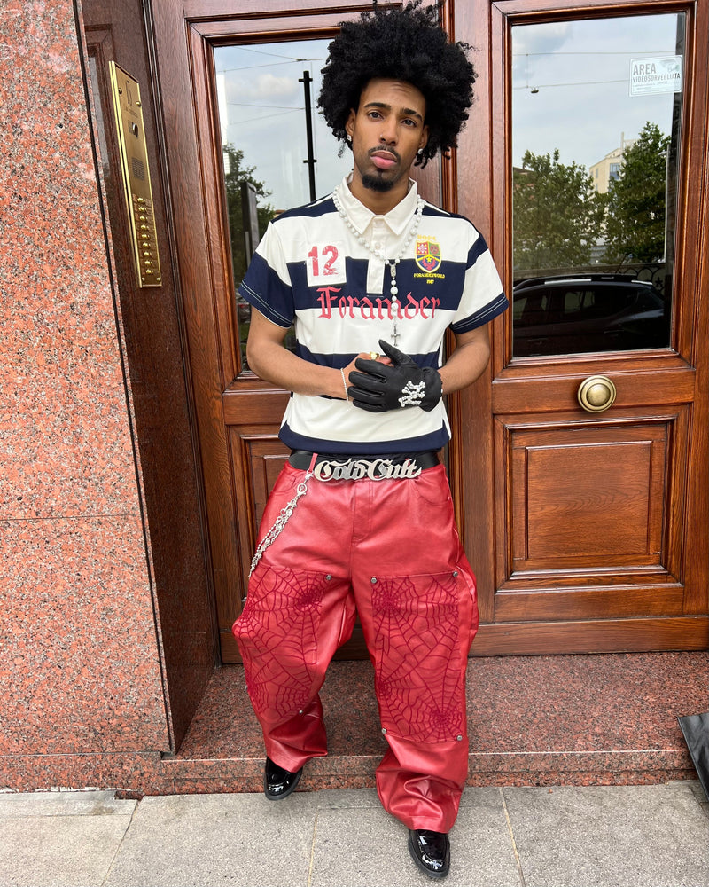 Model wearing a navy and white striped rugby polo with crest detail, styled with red leather pants and gloves outside a doorway