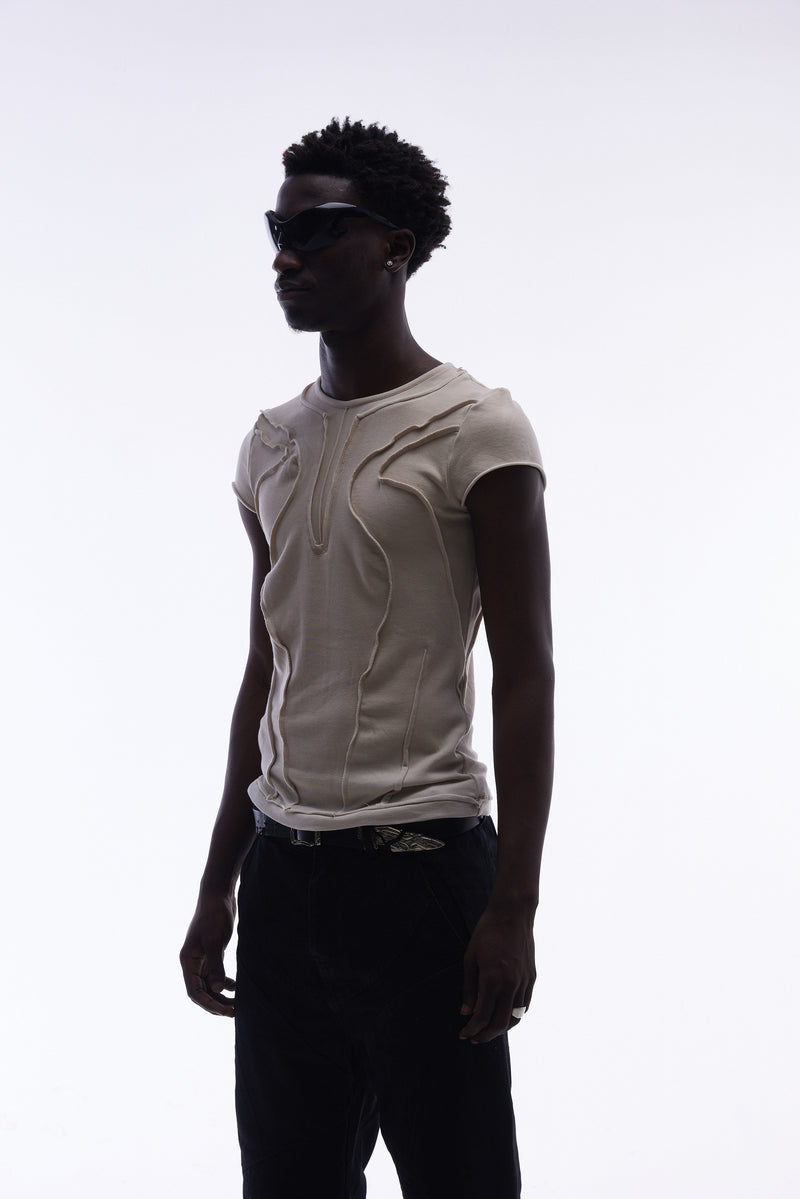 Beige sculpted cotton tee with raw seam paneling, worn by a model in black sunglasses and dark pants