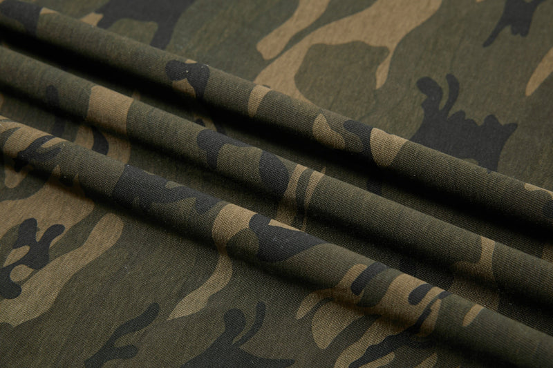 Close-up of olive and tan camo fabric with folded texture, featuring a bold streetwear graphic T-shirt pattern