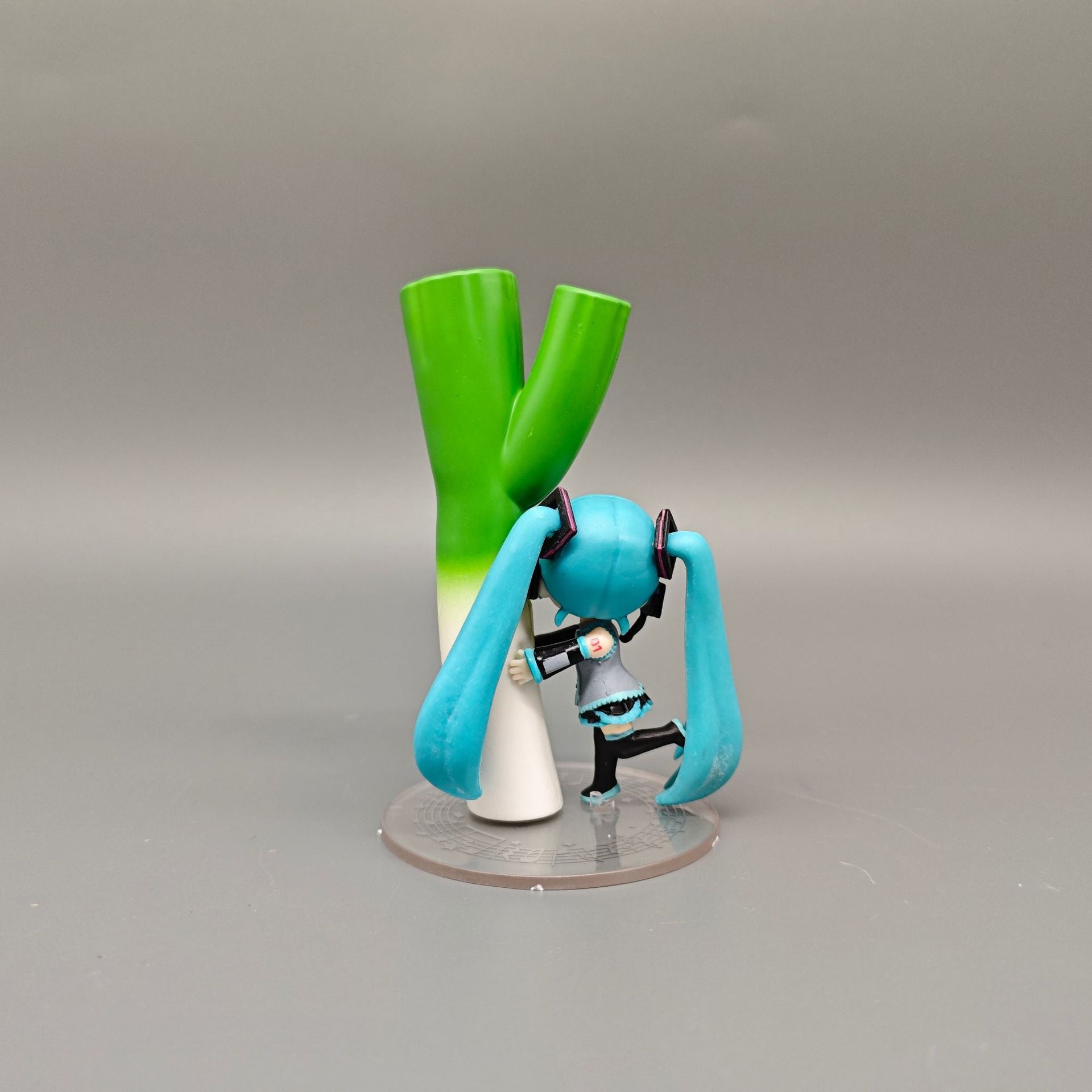 The Chibi Hatsune Miku Holding Green Onion 12cm PVC Figure features a playful Q-version of Miku with long teal hair, dressed in black and teal, holding a large green onion on a round base against a plain gray background.