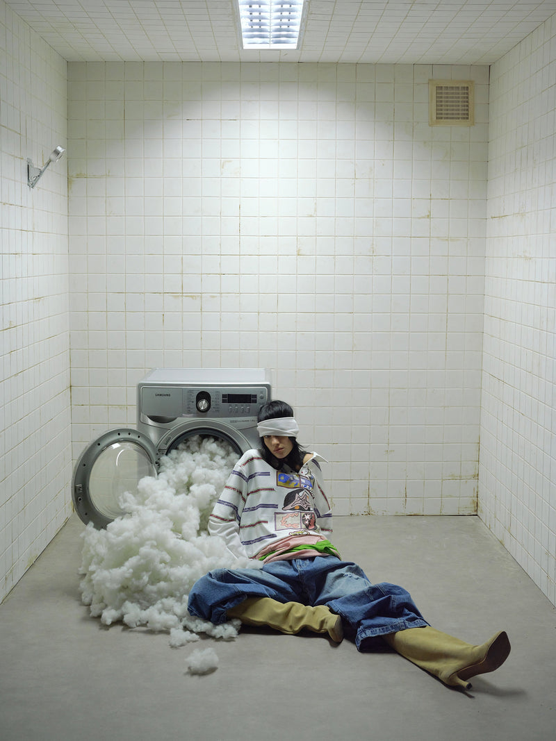 Model wearing white graphic long-sleeve polo shirt sitting beside open washing machine