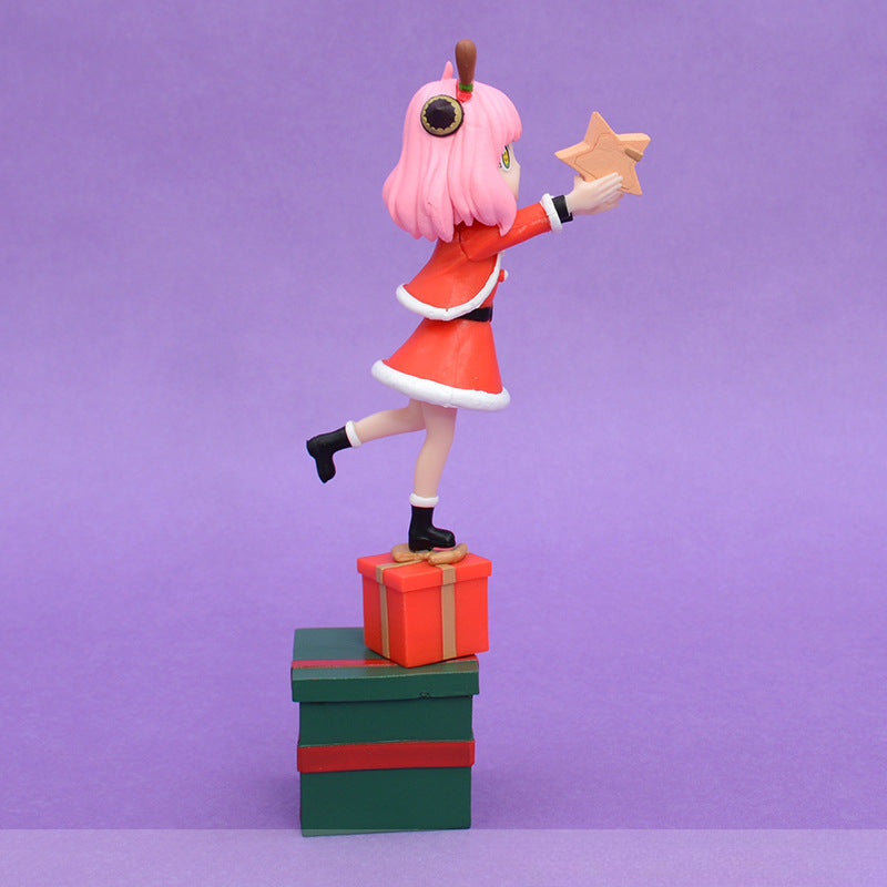 The Anya Forger Christmas PVC Figure - 18cm Holiday Edition from Spy × Family features a pink-haired girl in a festive red and white outfit. On tiptoe atop red and green gift boxes against a purple background, she reaches forward with a wooden star.