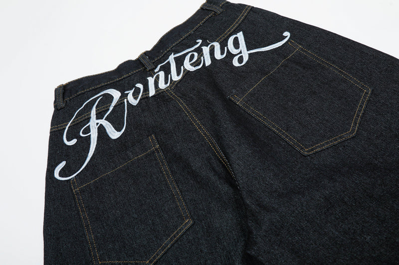 Back waist close-up of black denim jeans with white script embroidery and rear pockets