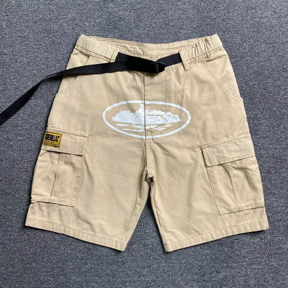 Beige Corteiz cargo shorts laid flat, showing white oval front graphic print, dual cargo pockets, and black webbing belt with buckle