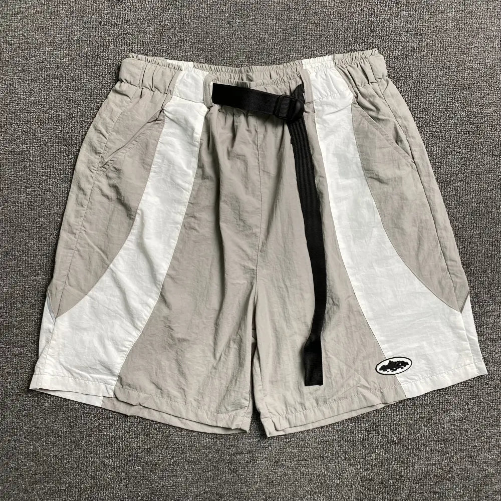 Corteiz beige and white colorblock nylon shorts laid flat, showing curved panel design, black webbing belt with buckle, and small oval logo patch on the lower leg