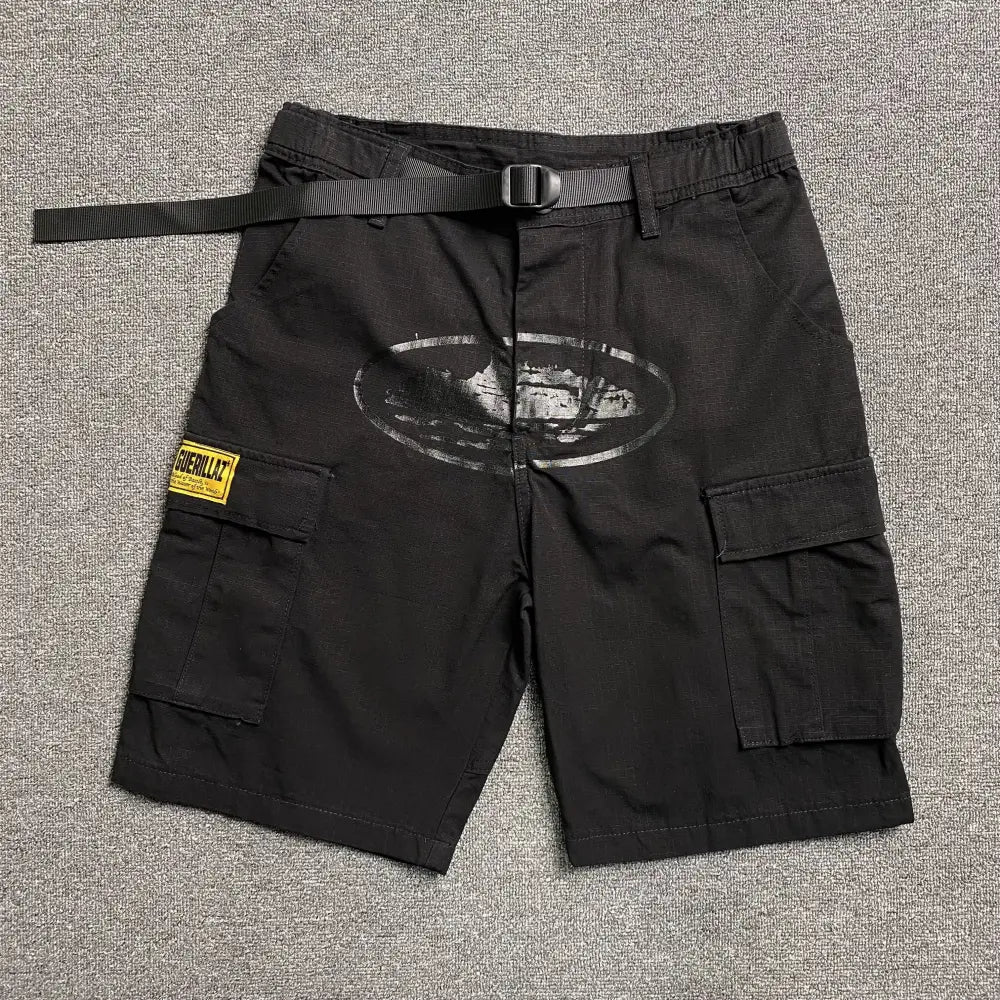 Black Corteiz cargo shorts laid flat, showing a front oval graphic print, Guerillaz woven patch on the left cargo pocket, and an attached nylon webbing belt with buckle.