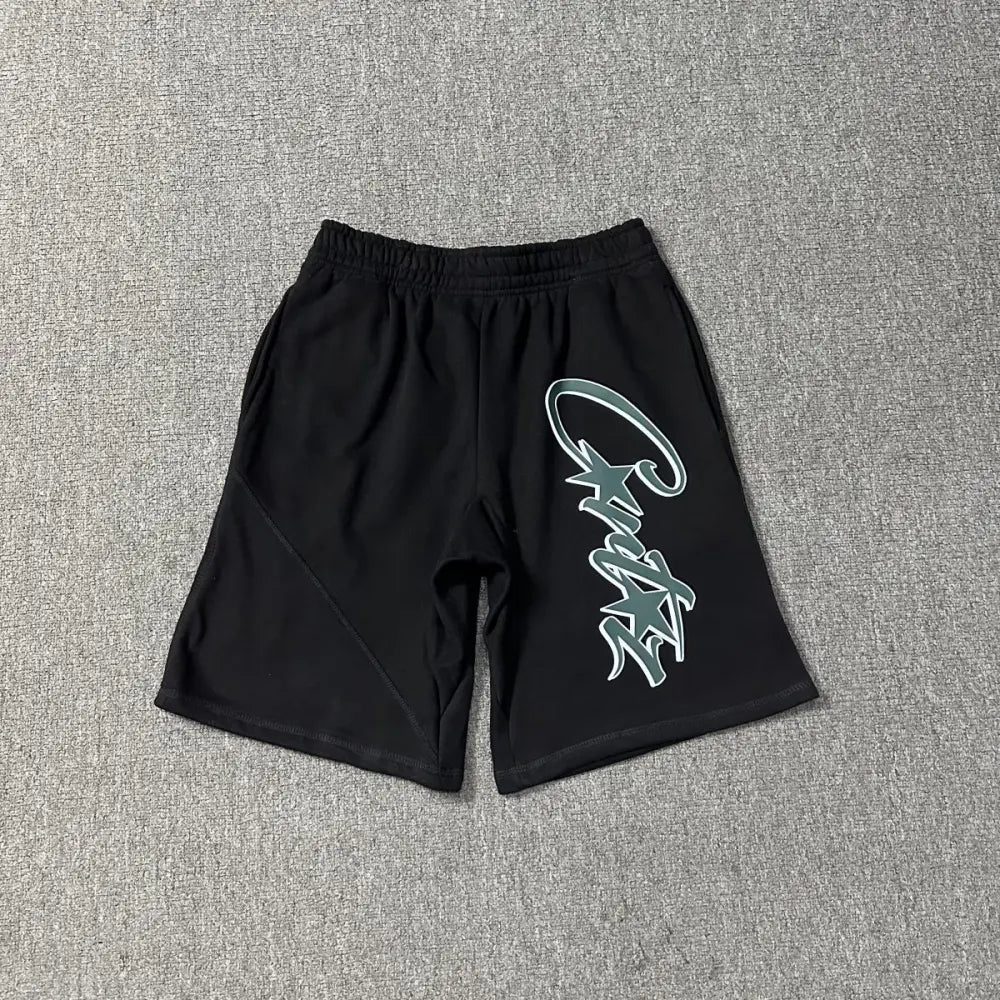 Black Corteiz jogger shorts laid flat, showing a green and white graffiti-style letter print on the front left leg and elasticated waistband