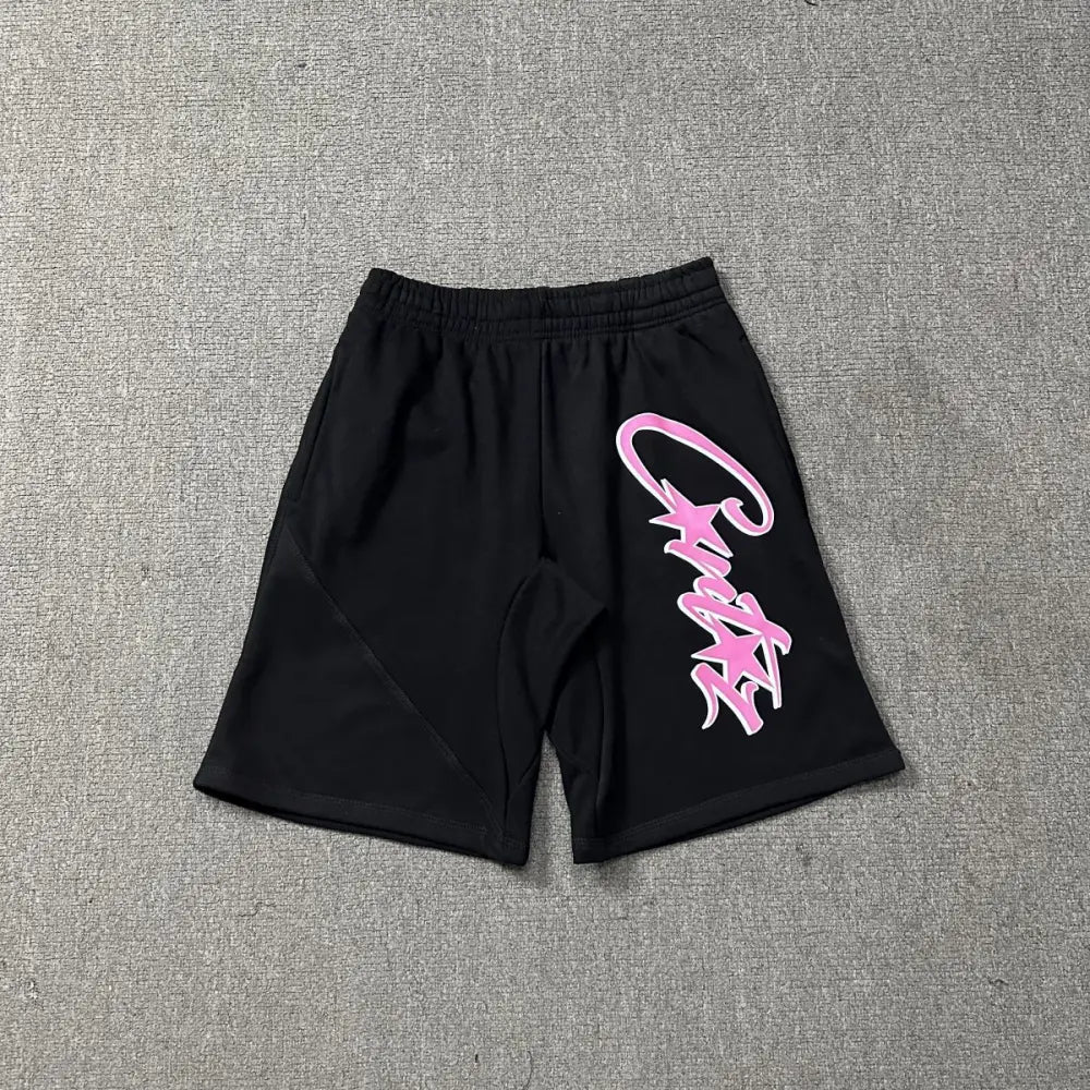 Black cotton jogger shorts laid flat, featuring a large pink graffiti-style letter print across the front leg and an elastic waistband