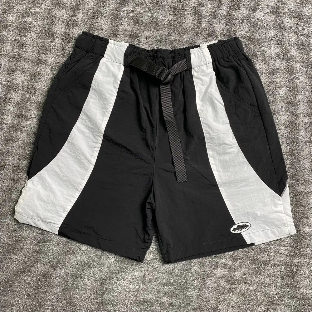 Corteiz black and white colorblock nylon shorts laid flat, showing webbing belt buckle and oval logo patch on the right leg