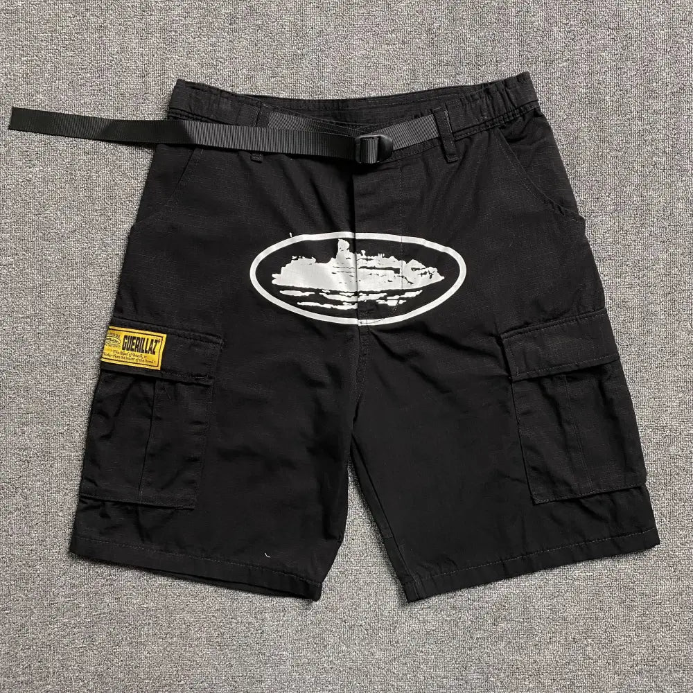 Black Corteiz cargo shorts laid flat, showing a white oval graphic print on the front, multiple cargo pockets, and an attached webbing belt with buckle