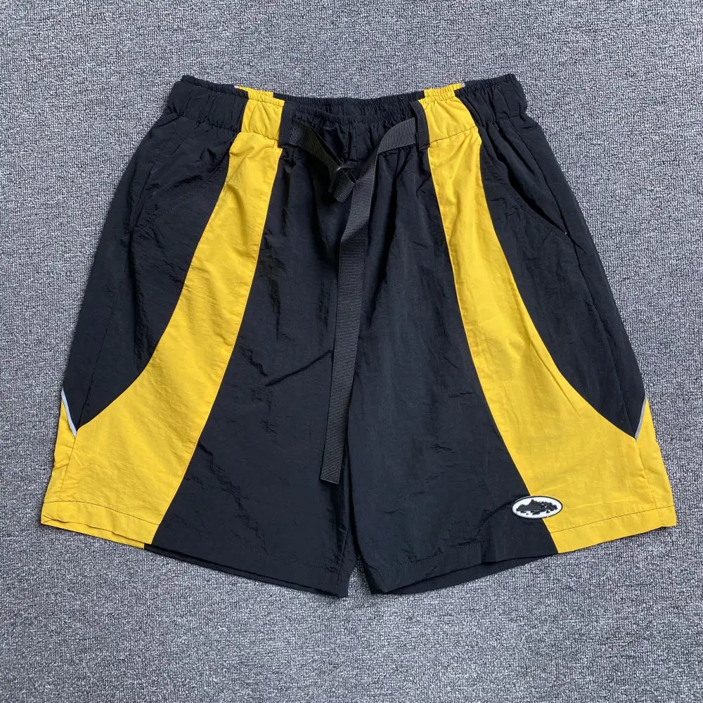 Corteiz black and yellow colorblock nylon shorts laid flat, showing curved yellow side panels, drawstring waistband with woven strap, and oval logo badge on front hem