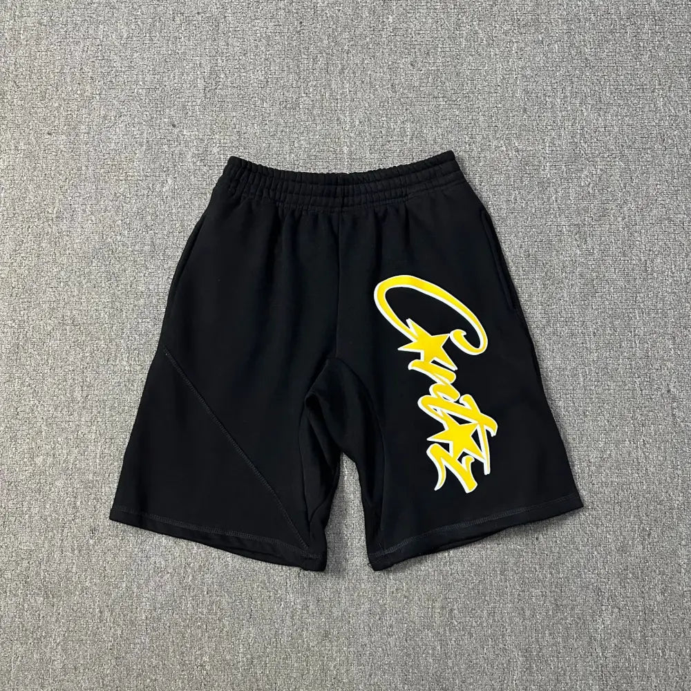 Black cotton Corteiz jogger shorts laid flat, showing a large yellow and white script letter print on the front leg