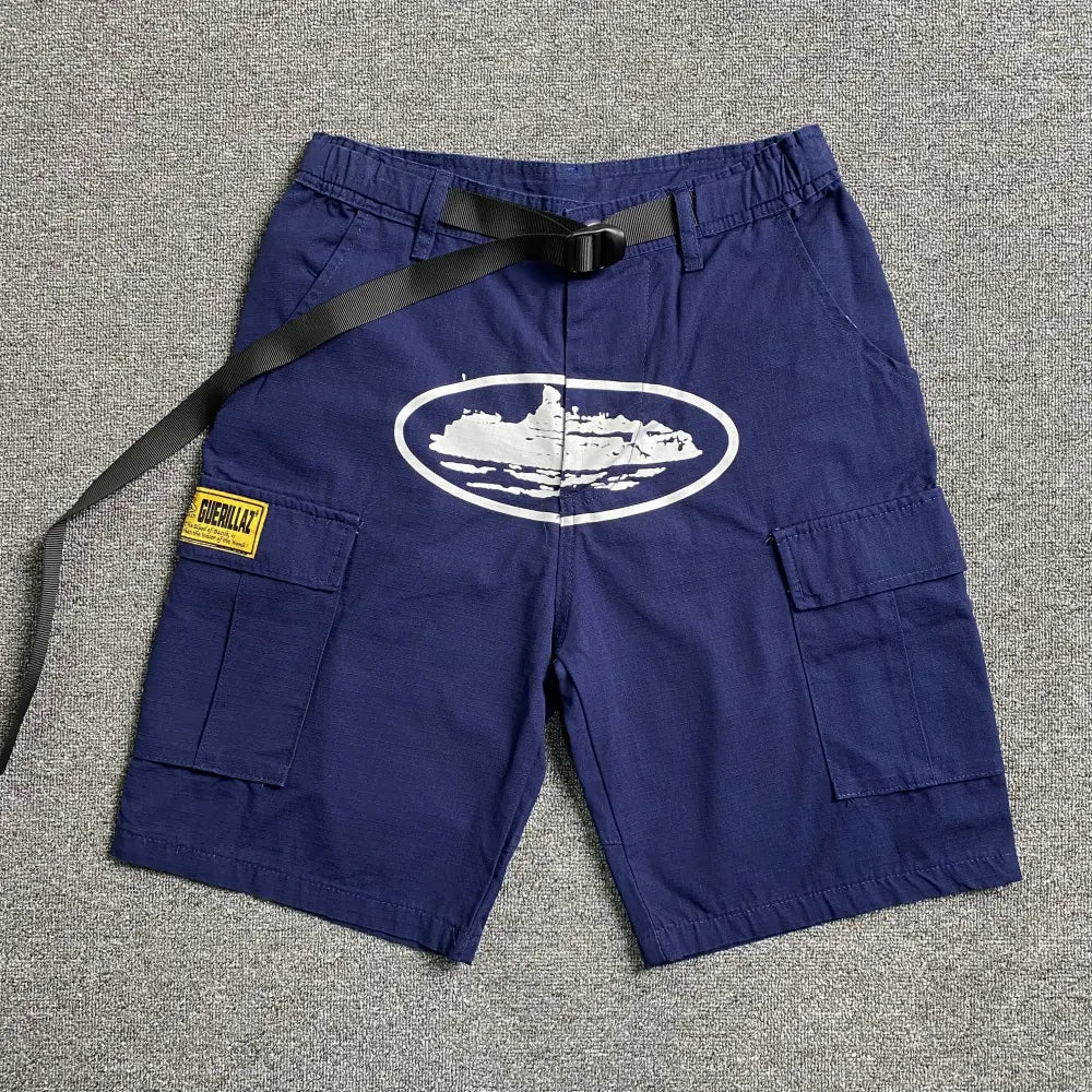 Navy blue Corteiz cargo shorts laid flat, showing a white oval graphic print on the front, cargo pockets with flap closures, a woven Guerillaz patch on the left pocket, and an adju