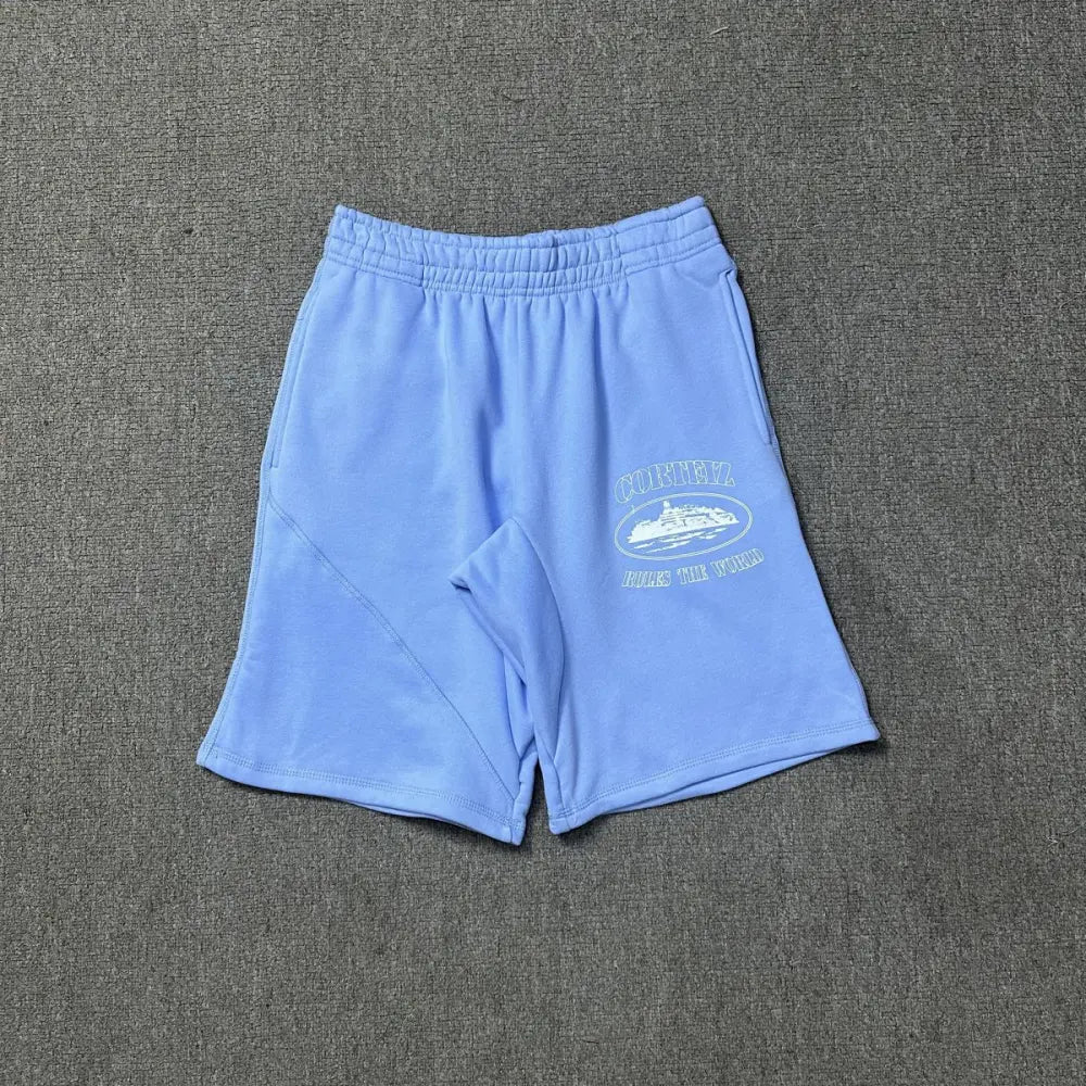 Light blue Corteiz cotton jogger shorts laid flat, showing the front 'Corteiz Rules The World' oval graphic print with yacht illustration