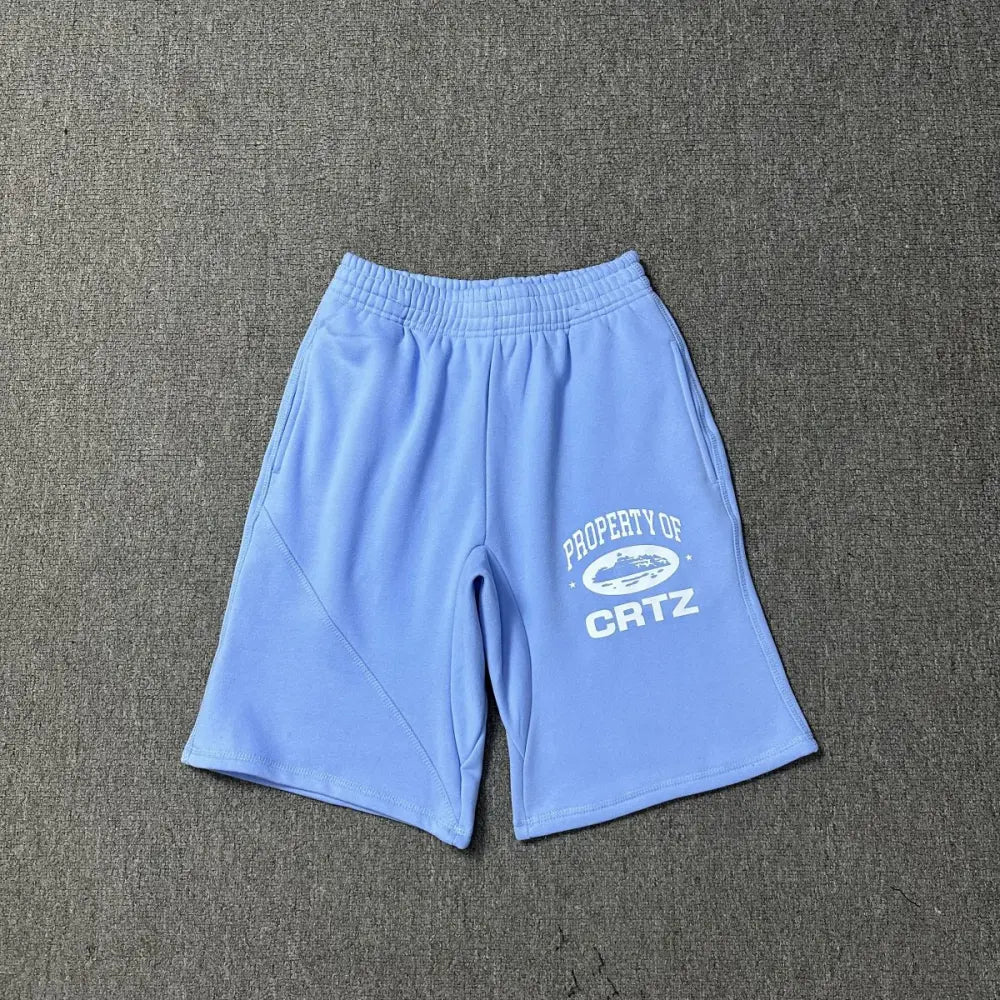 Light blue Corteiz cotton jogger shorts laid flat, showing white Property of CRTZ graphic print on the left leg and elasticated waistband