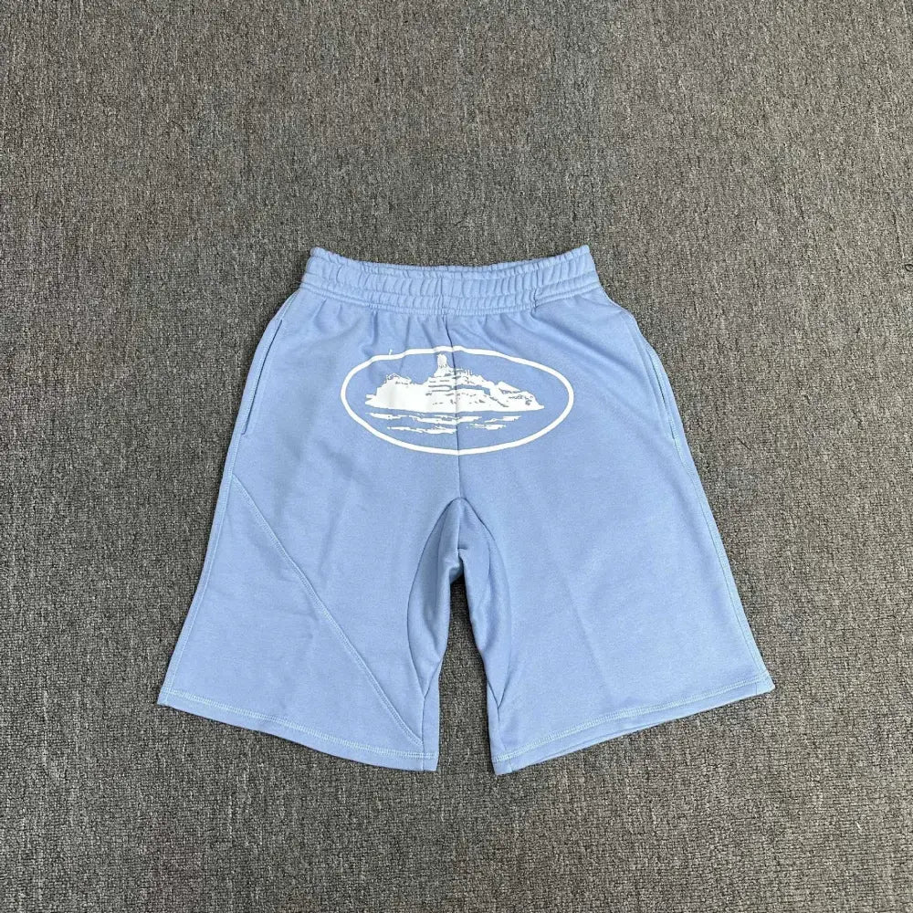 Light blue Corteiz cotton shorts laid flat, showing a large white oval logo graphic on the front panel and elasticated waistband
