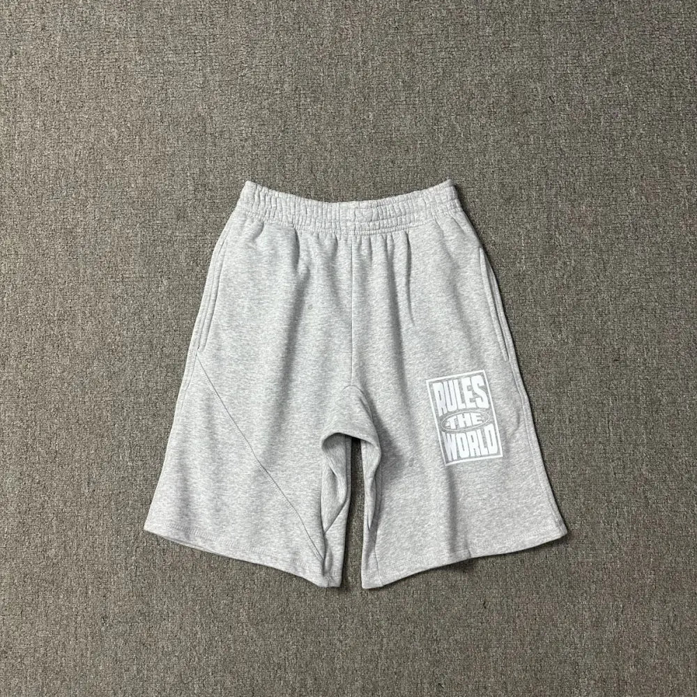 Light grey Corteiz cotton jogger shorts laid flat, showing a white Rules The World graphic print on the left leg and an elasticated drawstring waistband