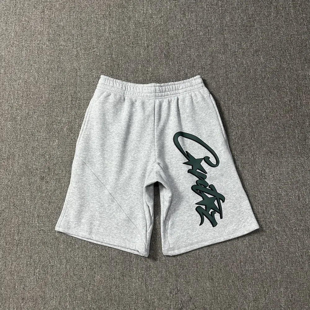 Light grey Corteiz cotton shorts laid flat, showing a large dark green script logo print on the front leg