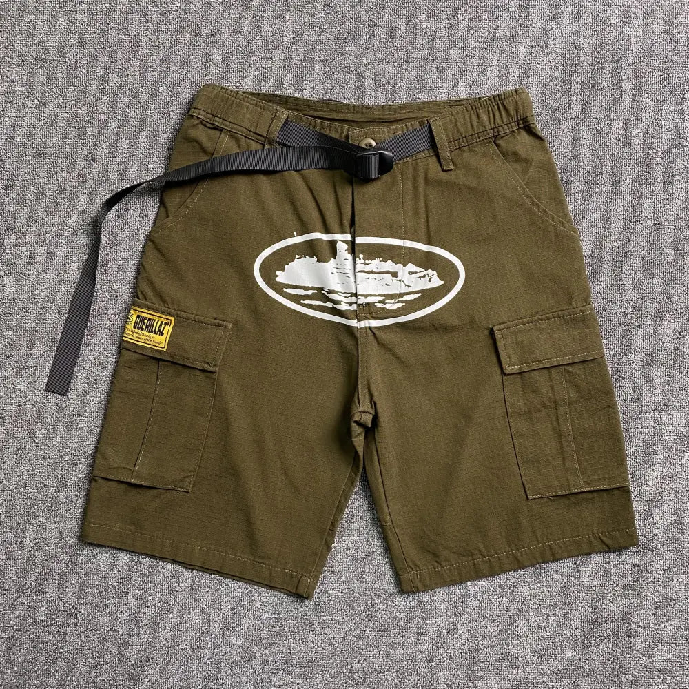Olive green Corteiz cargo shorts laid flat, showing white oval front graphic, buckle belt, and cargo pockets