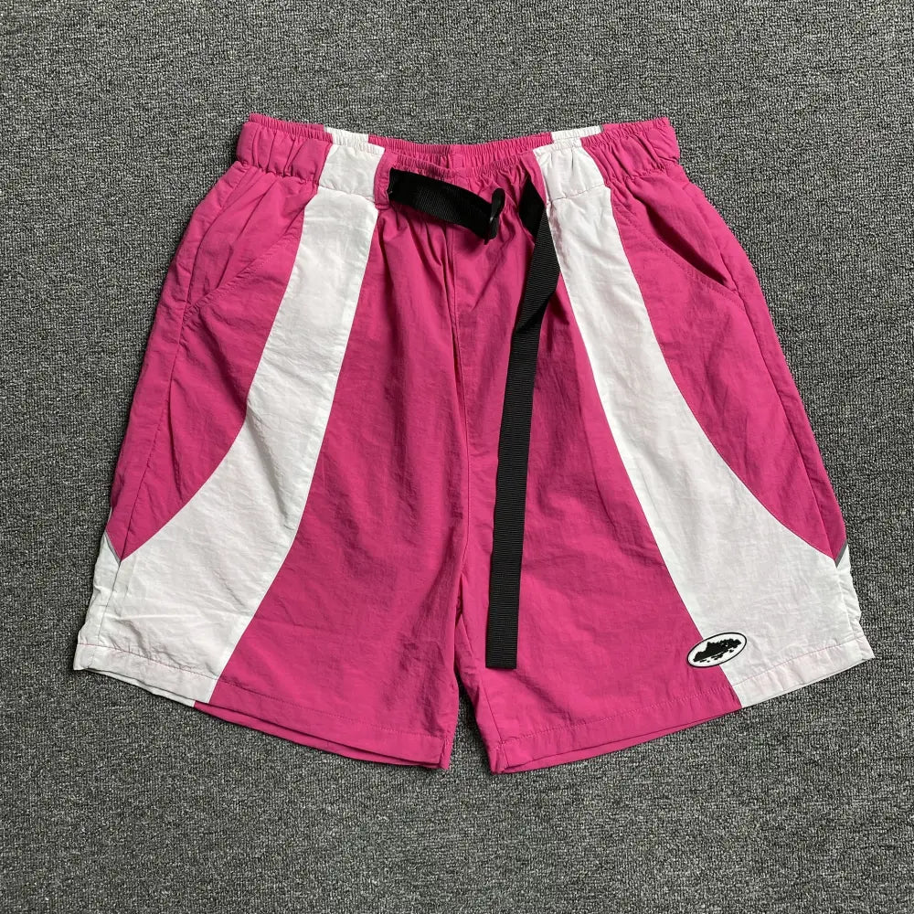 Pink Corteiz nylon shorts laid flat, showing white colour block panels, black clip buckle waistband, and logo patch on the lower leg