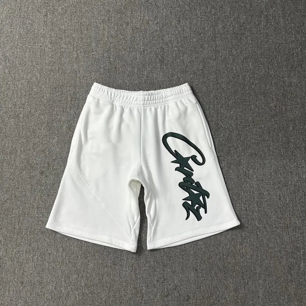 White cotton Corteiz jogger shorts laid flat, showing a large dark green script logo graphic printed down the right leg