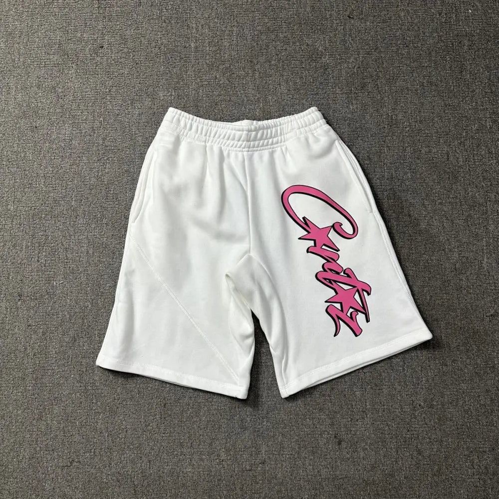 White cotton Corteiz jogger shorts laid flat, showing a large pink script logo print on the front leg