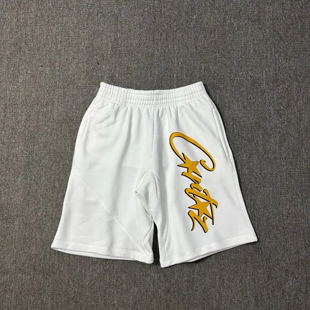 White cotton jogger shorts laid flat, showing a large yellow script letter print with star details on the left leg