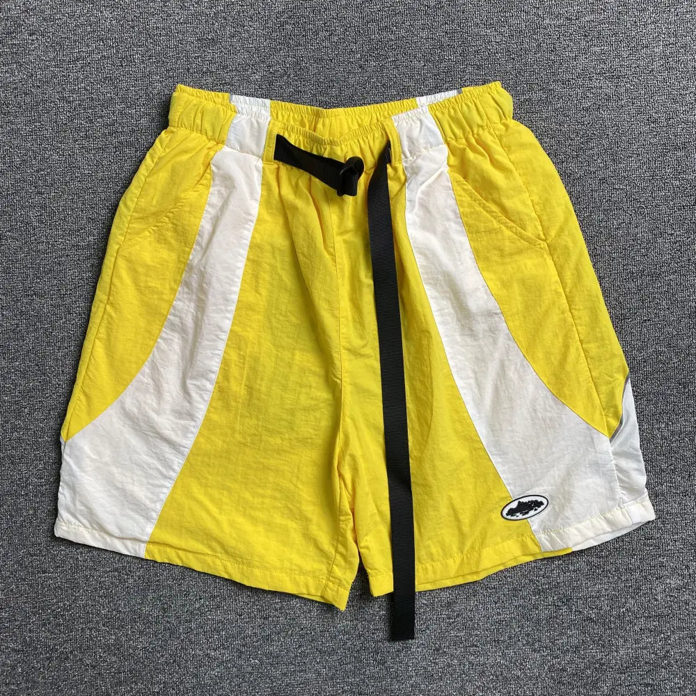 Corteiz yellow and white colorblock nylon shorts laid flat, showing curved panel design, black buckle strap, and embroidered logo patch