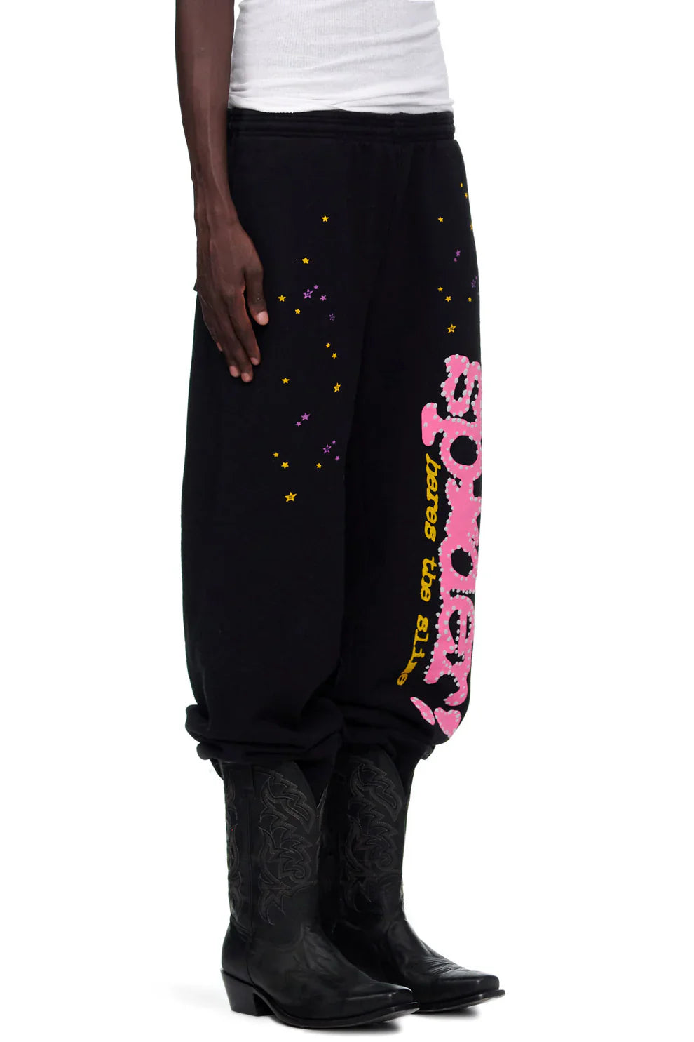A person wears Sp5der Sweatpants – unisex black cotton with pink graphic logo and star print – slightly rolled at the cuffs, paired with black cowboy boots and a white top for a laid-back look.