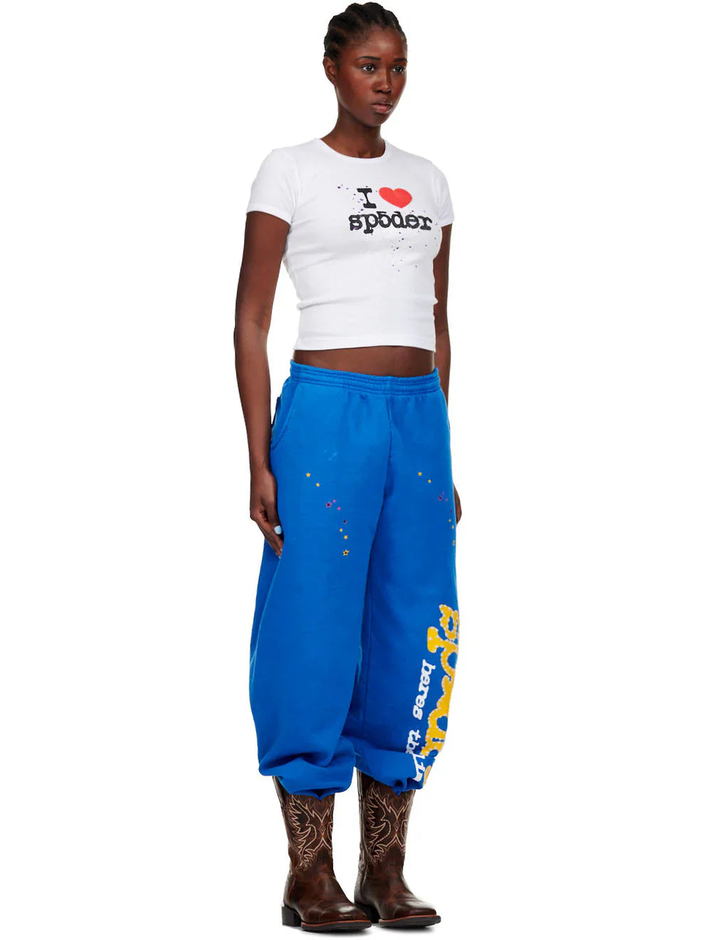 A woman with cornrows faces forward, wearing a white I ♥ sp8der cropped t-shirt, Sp5der Sweatpants – unisex blue cotton sweatpants with a yellow side logo and star print, and brown cowboy boots.