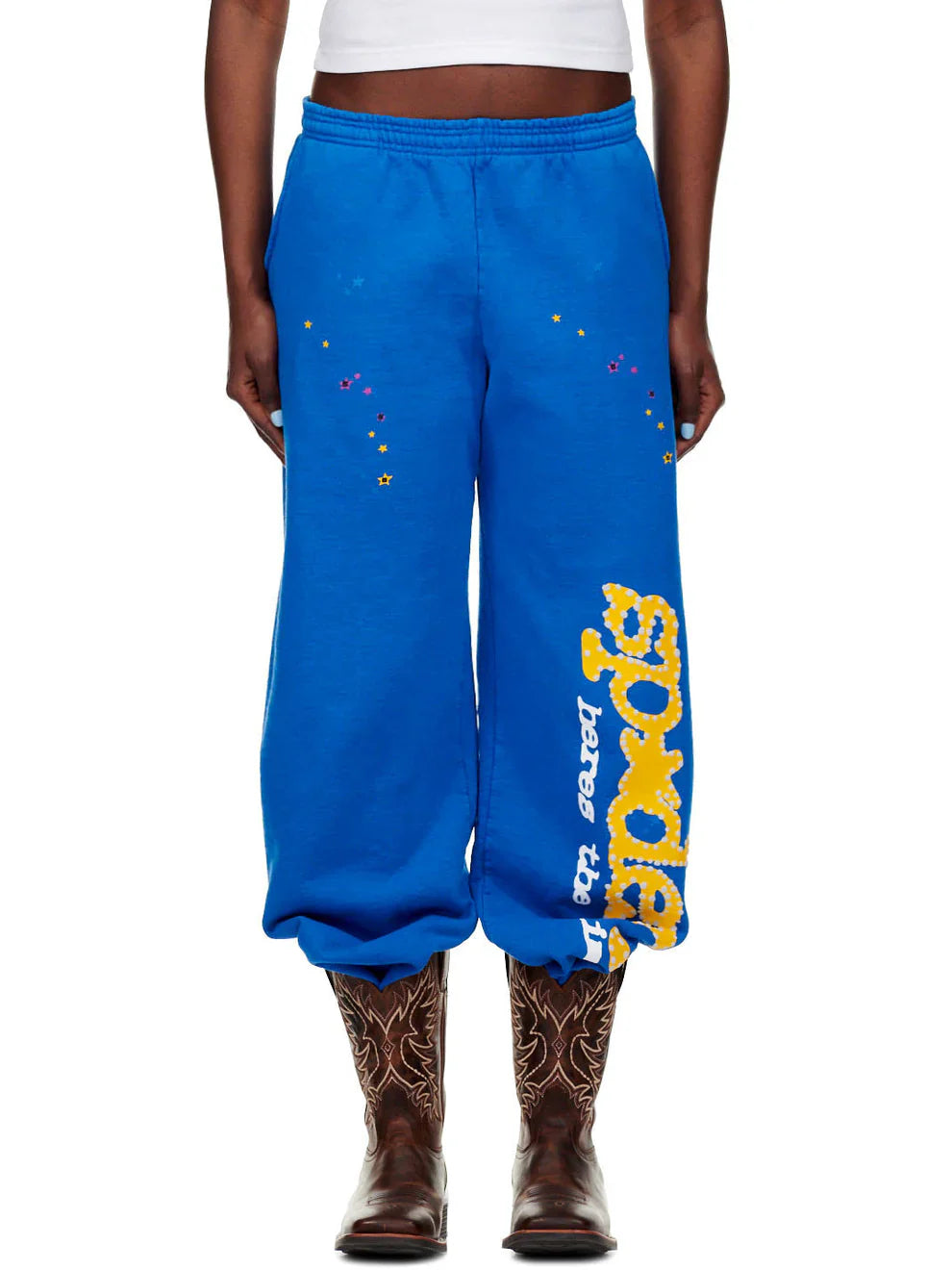 Someone is dressed in Sp5der Sweatpants – unisex blue cotton sweatpants with a yellow side logo and star print, paired with a white crop top and brown cowboy boots.