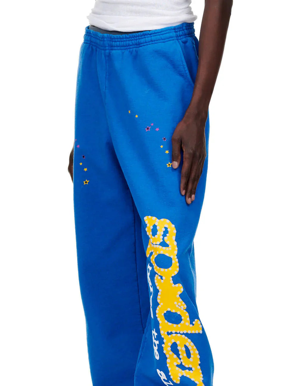 Only the lower torso and hand are shown wearing Sp5der Sweatpants—Unisex Blue Cotton Sweatpants featuring a yellow side logo and bold star prints, including star accents near the hip for a standout look.