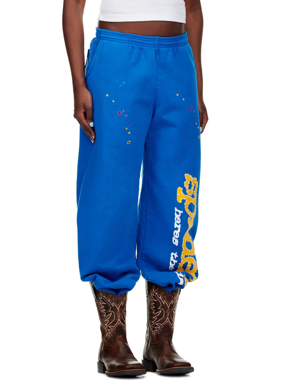 A person wearing Sp5der Sweatpants – unisex blue cotton sweatpants with a yellow side logo and star print, paired with a white cropped top and brown embroidered cowboy boots.