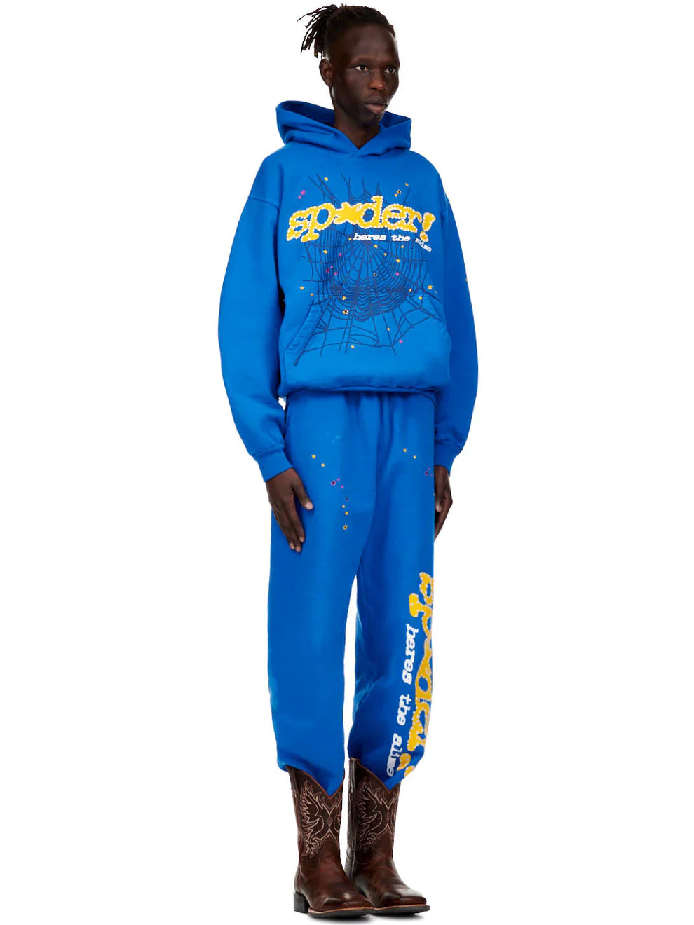 A person wears Sp5der Sweatpants—unisex blue cotton with yellow side logo and star print—plus a matching hoodie and brown cowboy boots, blending streetwear joggers with western flair against a white background.