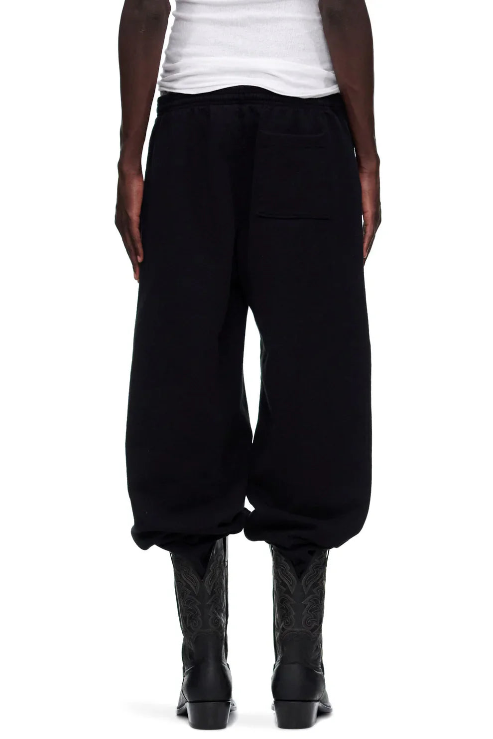 A person in a white tank top, Sp5der Unisex Black Cotton Sweatpants with Pink Graphic Logo and Star Print, and black patterned boots stands facing away from the camera against a plain background.