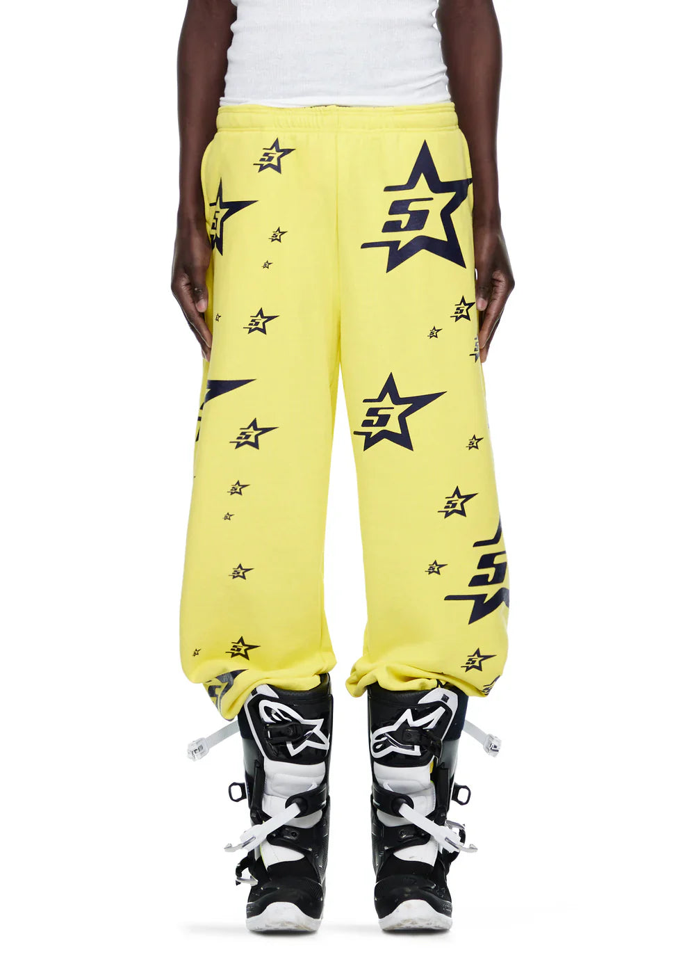 A person wears Sp5der Sweatpants – unisex yellow cotton sweatpants with all-over star print, paired with black and white motocross boots. Their hands are by their sides and the upper body is partially visible.