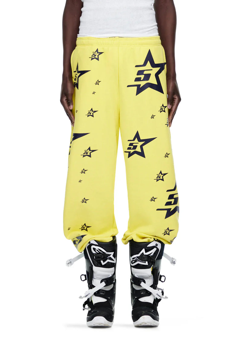 A person wears Sp5der Sweatpants – unisex yellow cotton sweatpants with all-over star print, paired with black and white motocross boots. Their hands are by their sides and the upper body is partially visible.