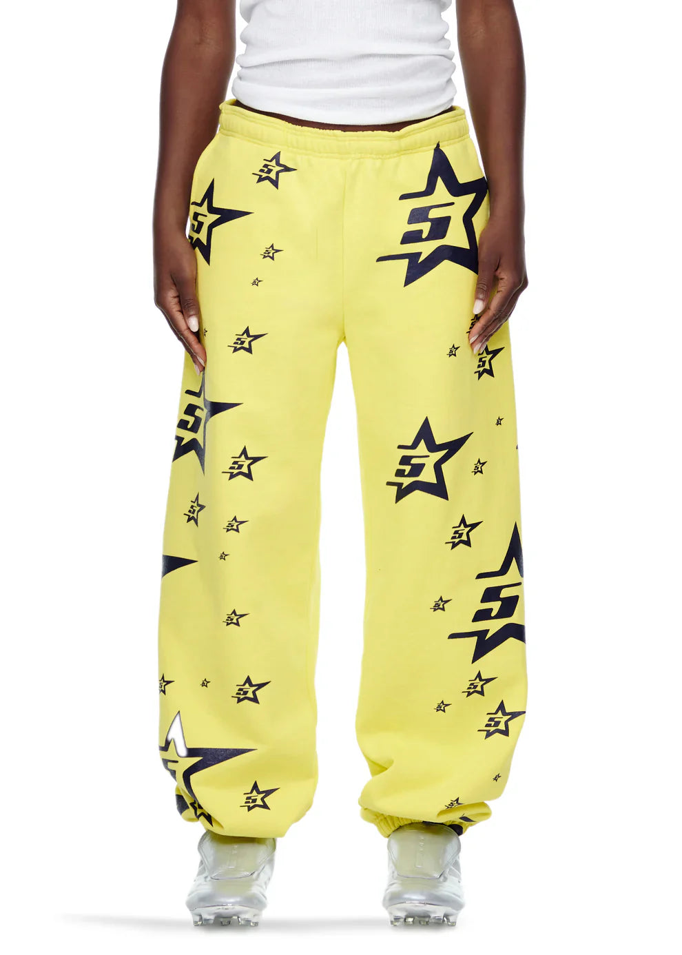 Someone dressed in Sp5der Sweatpants – unisex yellow cotton sweatpants with an all-over star print – paired with a white tank top and translucent sneakers.