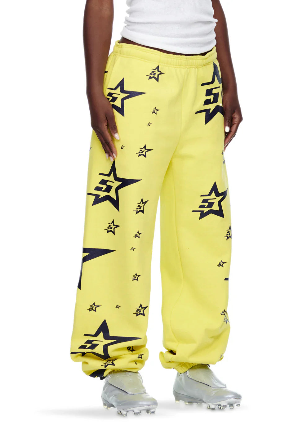 A person wearing unisex yellow Sp5der Sweatpants with all-over star print, a white tank top, and silver athletic shoes stands against a plain white background.