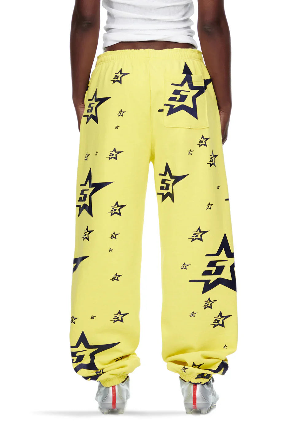 A person wears Sp5der Sweatpants—unisex yellow cotton sweatpants with an all-over star print—paired with white sneakers and a white crop top. The photo, taken from behind, shows the full length of the pants.