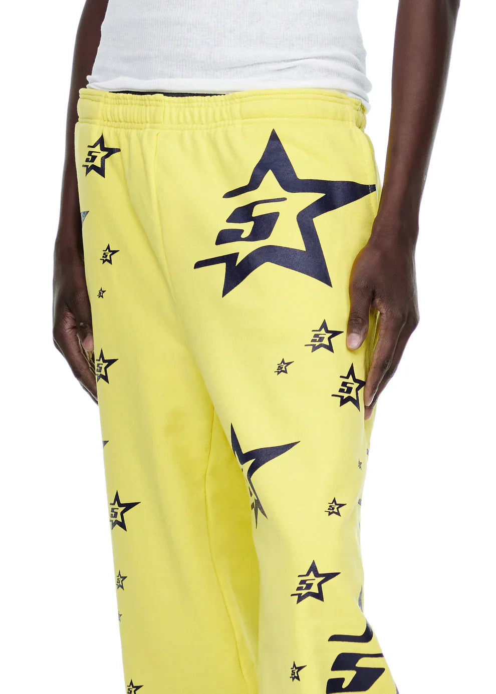 A person wears Sp5der Sweatpants – Unisex Yellow Cotton Sweatpants with All-Over Star Print and a white tank top. Only the lower torso, right arm, and part of the legs are visible.