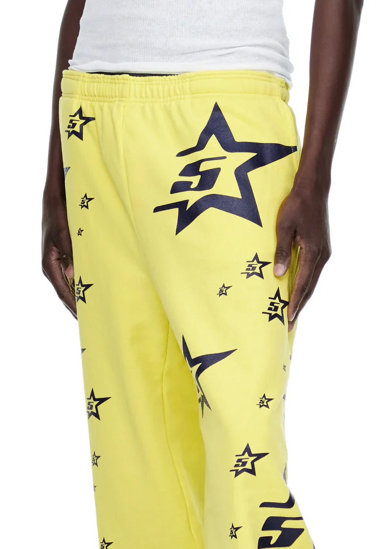 A person wears Sp5der Sweatpants – Unisex Yellow Cotton Sweatpants with All-Over Star Print and a white tank top. Only the lower torso, right arm, and part of the legs are visible.