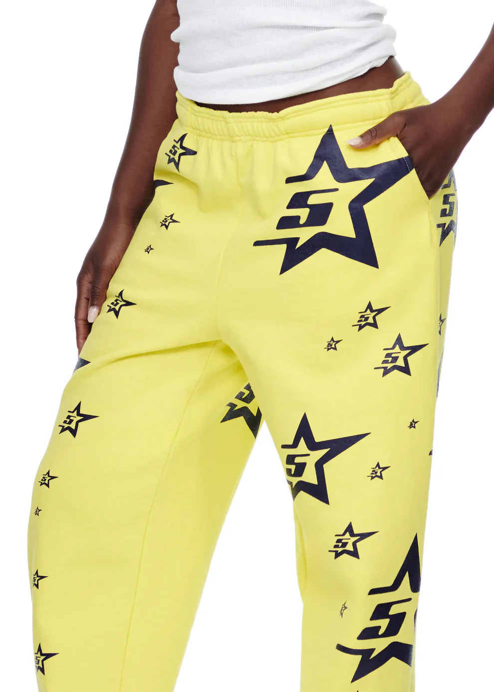 A person wears Sp5der Sweatpants – Unisex Yellow Cotton Sweatpants with an all-over star print, paired with a white sleeveless top. The image shows the lower torso and thighs, with one hand tucked into a pocket.