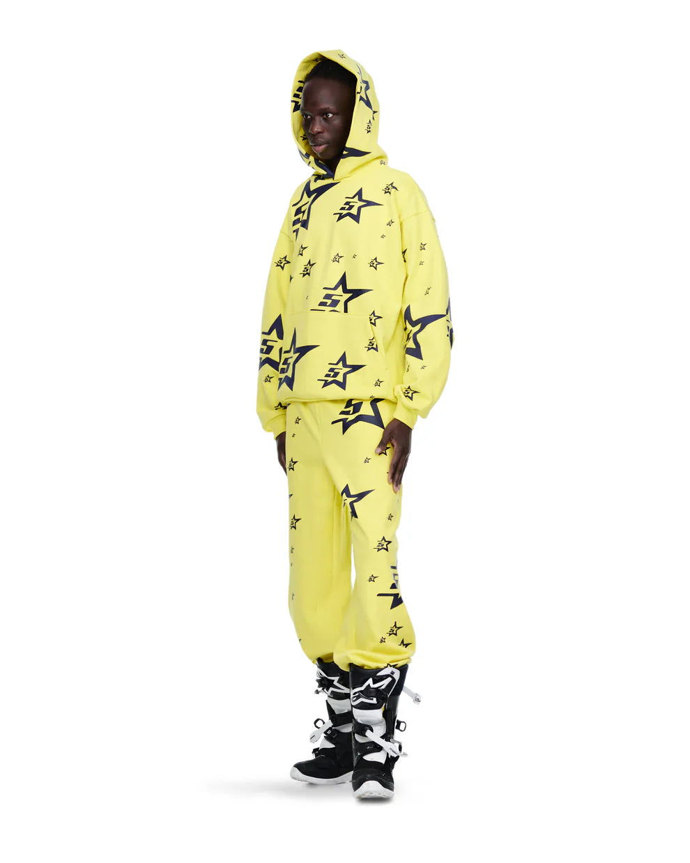 A person wears Sp5der Sweatpants – unisex yellow cotton sweatpants with an all-over star print – paired with black and white high-top sneakers and a raised hood, capturing a bold, urban streetwear vibe.