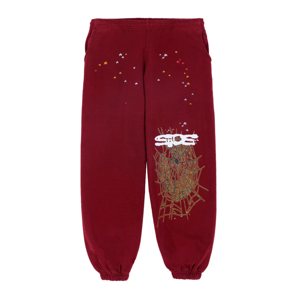 The Sp5der Sweatpants are unisex burgundy cotton sweatpants featuring elastic cuffs, small multicolored stars by the waist, and a bold spider web graphic with a spider and white SOS letters on the right leg.