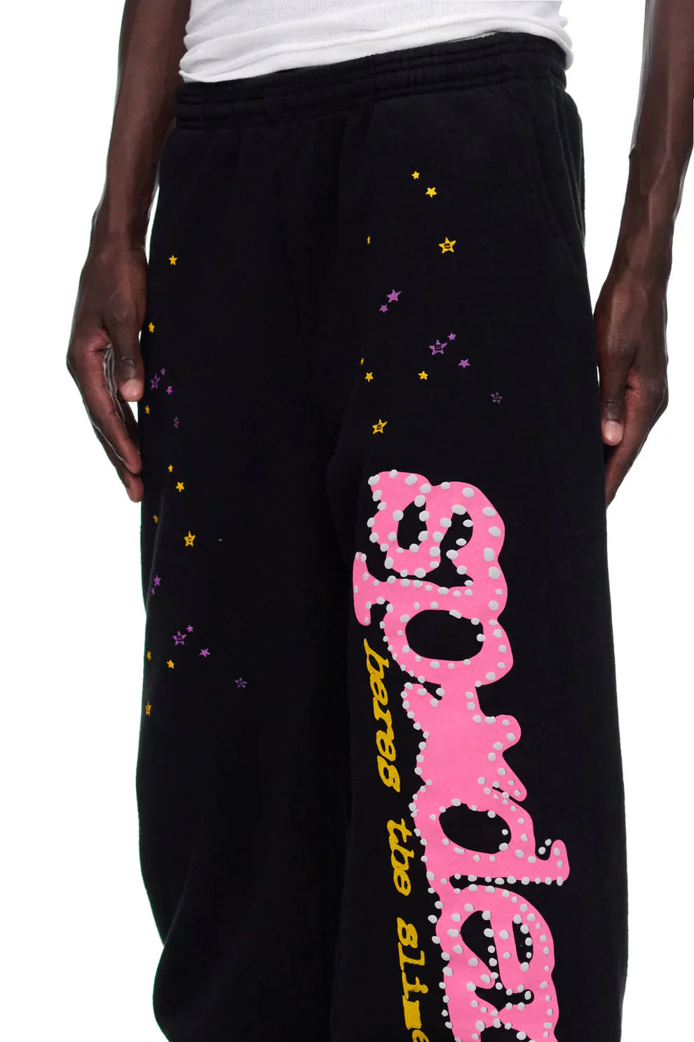 A person wearing Sp5der Sweatpants—unisex black cotton joggers featuring a pink graphic logo and colorful star print, perfect for bold streetwear style.