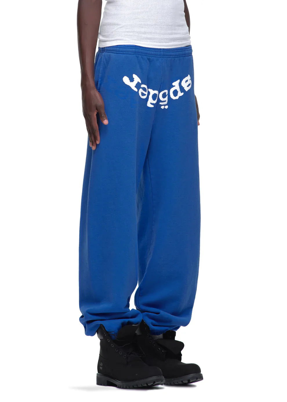 A person wears Sp5der Sweatpants – Unisex Blue Cotton Sweatpants with a front logo print, paired with a white tank top and black boots. Only the lower half is shown, highlighting these streetwear-inspired joggers.