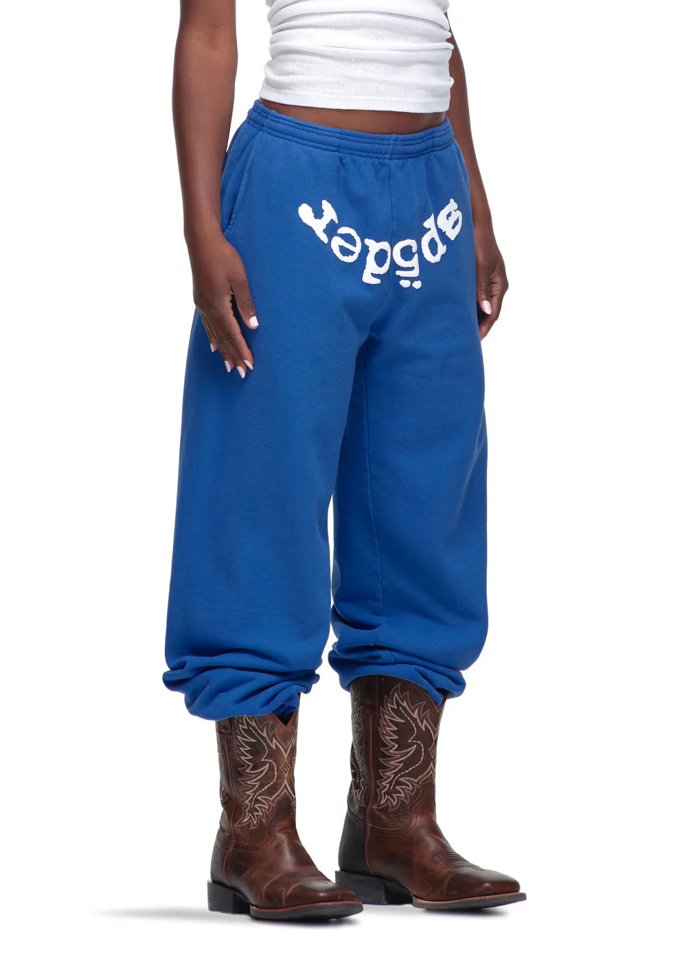 A person wears Sp5der Sweatpants – Unisex Blue Cotton Sweatpants with a front logo print, paired with a white sleeveless top and brown cowboy boots, standing against a white background to showcase bold streetwear style.